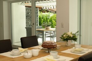 Uma sala de jantar com uma mesa posta para dois, um bolo em suporte de vidro e um vaso de planta.