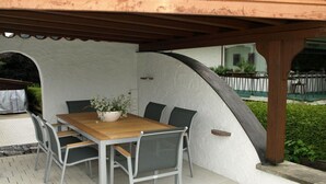 Une terrasse extérieure avec une table et des chaises en bois, une plante en pot et un mur blanc à la texture particulière.