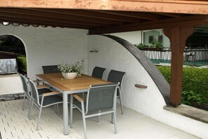 Um pátio externo com mesa e cadeiras de madeira, uma planta em vaso e uma parede branca com textura.