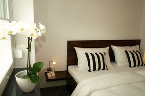Quarto de hotel com uma cama, almofadas brancas, mesa de cabeceira, abajur e uma orquídea em vaso.