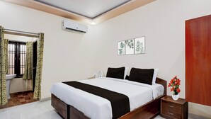 Quarto com cama de madeira, roupa de cama branca, mesa de cabeceira com um vaso de flores e ar-condicionado fixado na parede.