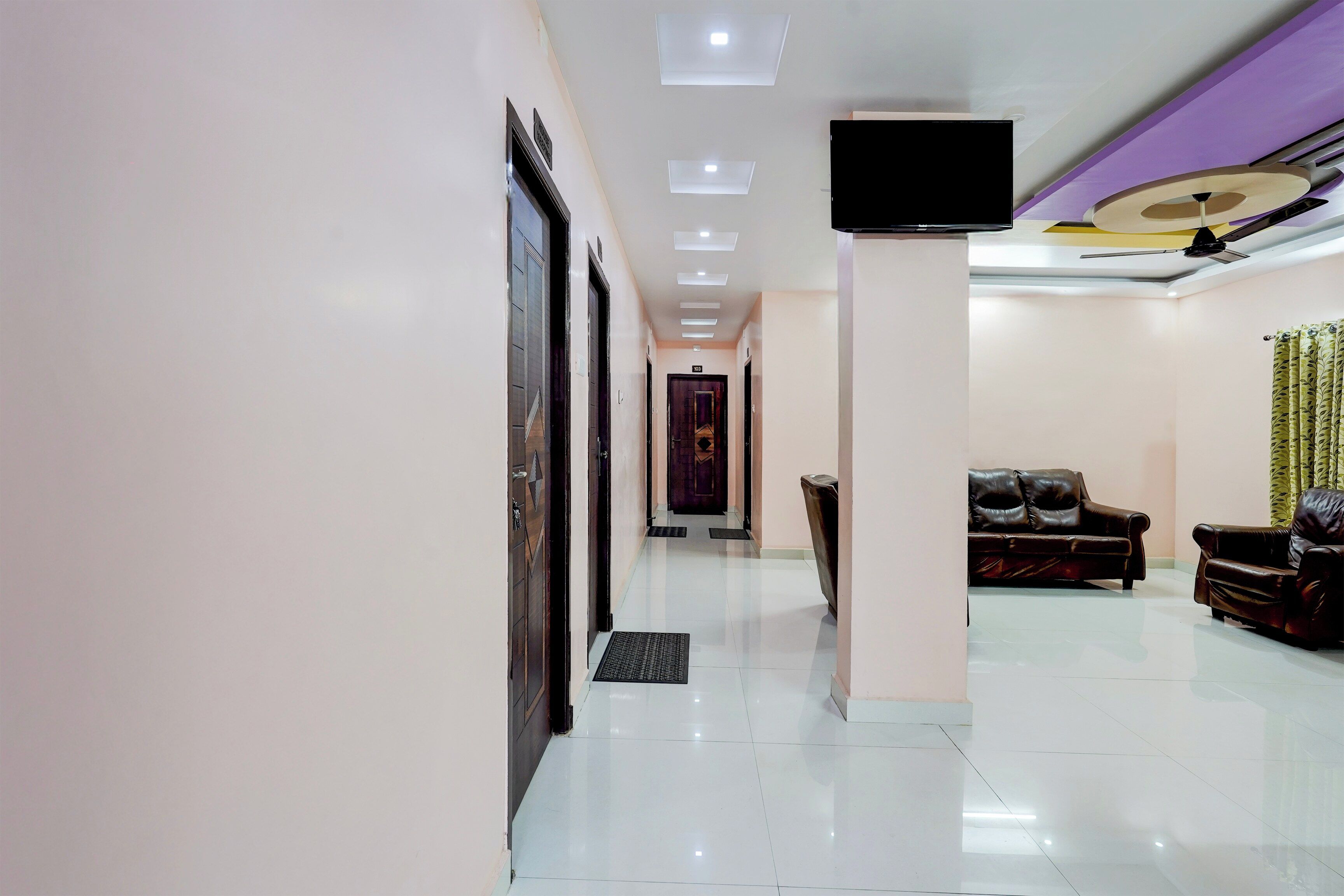 A hotel lobby with a reception desk, a flat-screen TV mounted on the wall, a seating area with leather sofas, and a tiled floor.