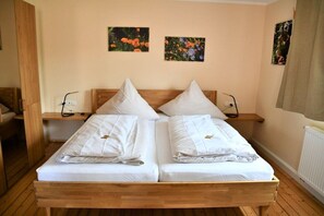 Ein Doppelbett mit weißer Bettwäsche, zwei Kissen und einem Holz-Kopfteil. Darüber hängen zwei Wandlampen und zwei gerahmte Blumenbilder.