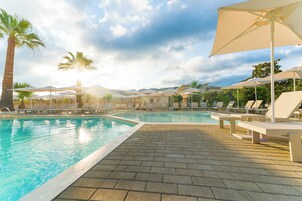 Una piscina con sillas de descanso y sombrillas, rodeada de palmeras y un cielo despejado.