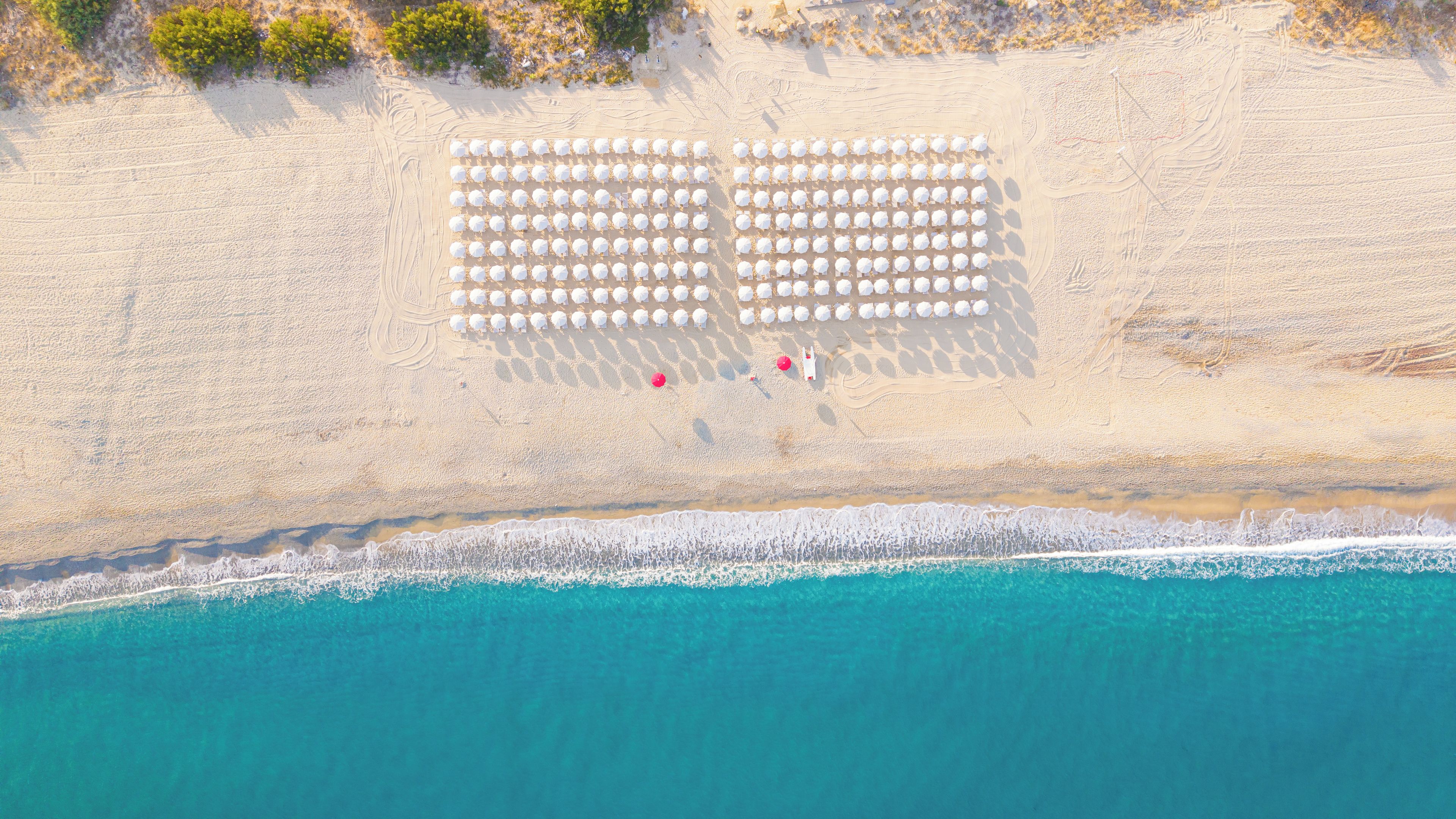 An aerial view of a beach with rows of white umbrellas and a few red chairs.