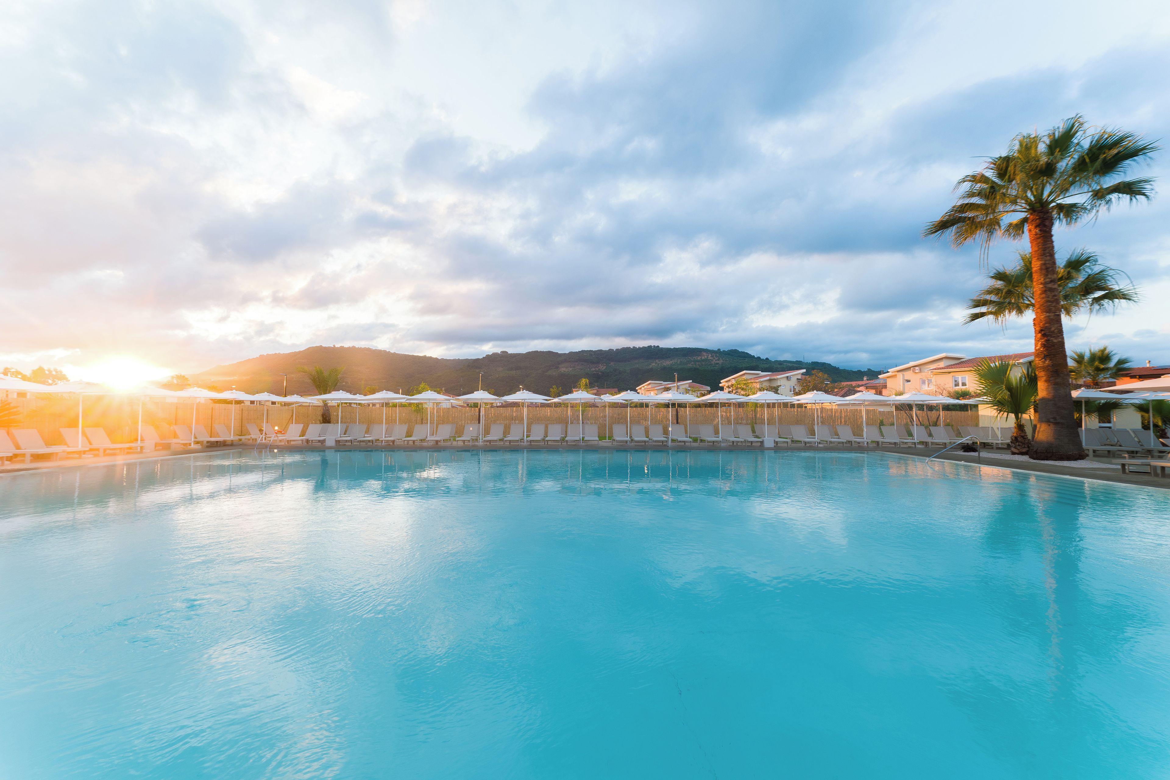 A swimming pool with lounge chairs and umbrellas, surrounded by a wooden fence and palm trees, with a mountain in the background.