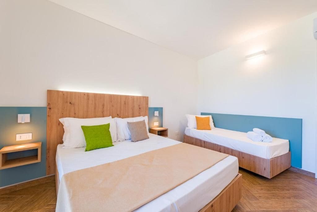 A hotel room with a large bed, a smaller bed, wooden headboards, bedside tables, and a wall-mounted light fixture.