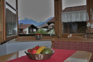 A room with a view of mountains through a window, a dining table with a red tablecloth, and a wicker basket with fruit. - Alpenflair holiday apartment 201 (Oberstdorf)