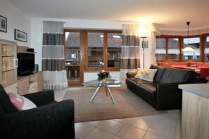 A living room with a sofa, a glass coffee table, a television, and a kitchen area visible through a glass door. - Alpenflair holiday apartment 201 (Oberstdorf)
