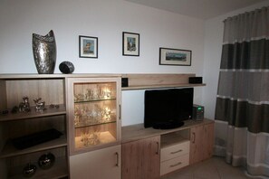 A living room with a TV on a wooden cabinet, a glass cabinet with wine glasses, and framed pictures on the wall. - Alpenflair holiday apartment 201 (Oberstdorf)
