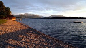 Une vue sur un lac, avec un quai, un bâtiment sur la gauche et des montagnes en arrière-plan.