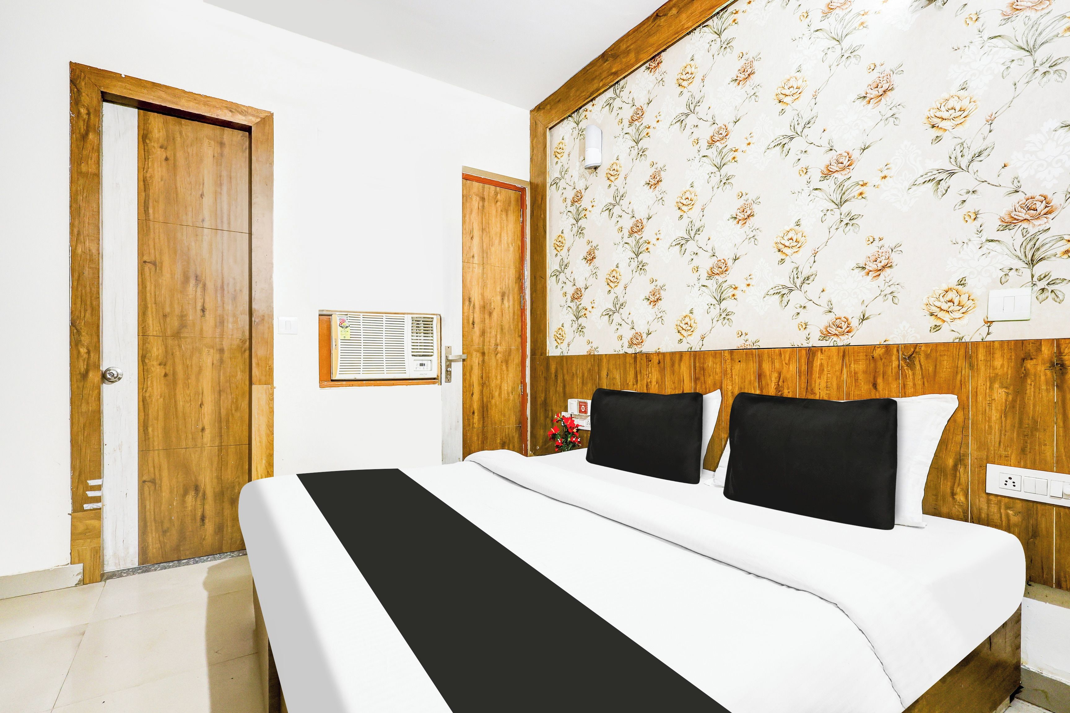 Ein Hotelzimmer mit einem hölzernen Kopfteil, einem Bett mit weißer Bettwäsche und schwarzen Kissen, einem blumigen Wandmuster und einer Holztür.