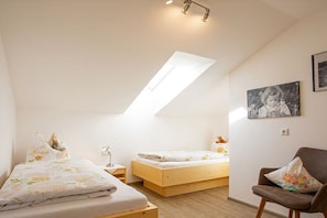 Quarto com teto inclinado, cama com roupa de cama floral, mesa de cabeceira, uma cadeira e um quadro na parede.