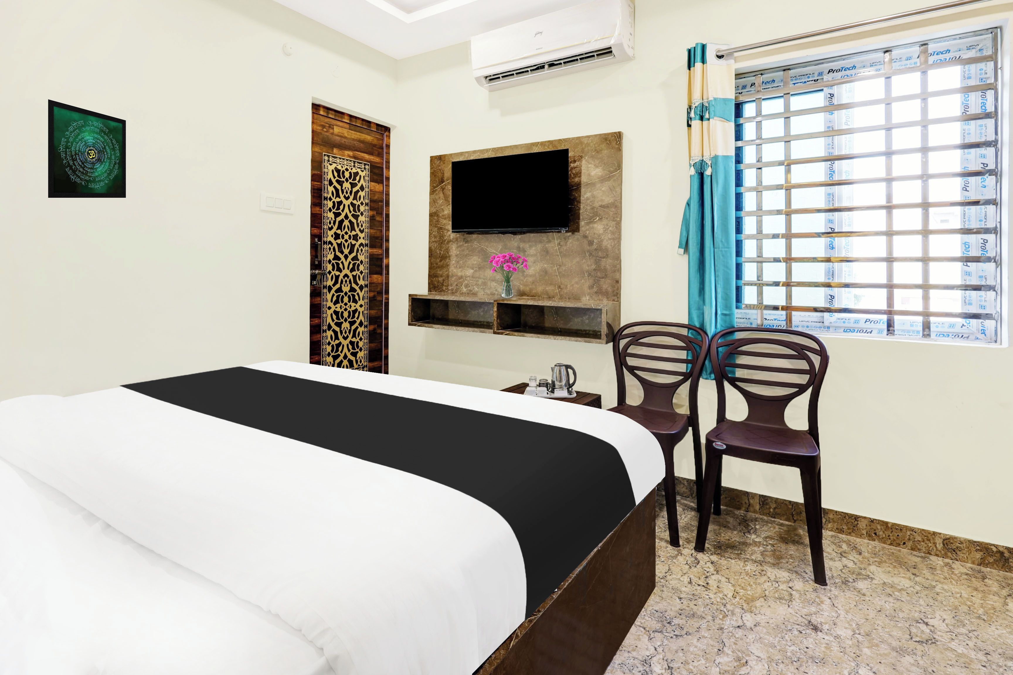 A hotel room with a bed, a flat-screen TV mounted on the wall, a small table with a vase of flowers, two chairs, and a window with curtains.