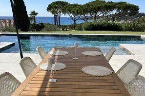 Una mesa de madera con individuales trenzados y sillas de mimbre junto a una piscina con vista al mar y a los árboles.