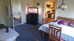 A room with a bed, a table with a chair, a television, and a kitchen area visible through an open door. - Holiday apartment with 1 bedroom (Wurster Nordseeküste)