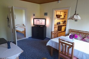 A room with a bed, a table with a chair, a television, and a kitchen area visible through an open door. - Holiday apartment with 1 bedroom (Wurster Nordseeküste)