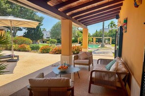 A covered outdoor seating area with wicker furniture, a round table with a bowl of fruit, and a view of a swimming pool and garden. - Villa in Cala Muarad with swimming pool (Cala Muarad)