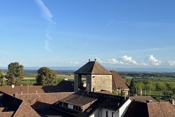 Um edifício com uma torre de pedra e um telhado de telhas vermelhas, rodeado por árvores e um céu claro.