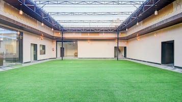 Un espacio interior con suelo de césped artificial verde, techo alto con estructura metálica y lámparas colgantes.
