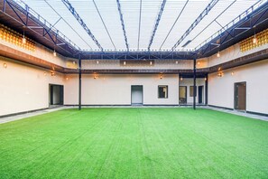 Un espacio interior con suelo de césped artificial verde, un techo alto con estructura de rejilla y varias puertas que dan a otras áreas.