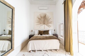A bedroom with a large mirror, a bed with a brown and white duvet, a wall-mounted air conditioner, and a window with a curtain. - Peaceful 5-Bedroom Riad in the Heart of Marrakech (Marrakesh)