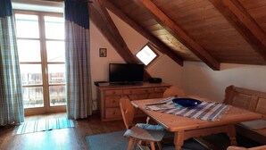 Acogedor cuarto ático con techo de madera, un televisor sobre un aparador, una mesa de comedor con sillas y un ventanal con cortinas.
