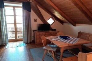 A cozy attic room with a wooden ceiling, a television on a dresser, a dining table with chairs, and a large window with curtains. - in the old forester's lodge (Riedlhütte)