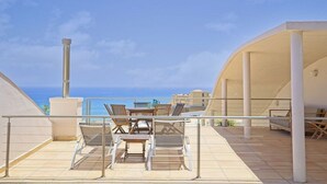 Una terrassa amb mobles d'exterior, una vista despejada del mar i un edifici amb teulada corbada.