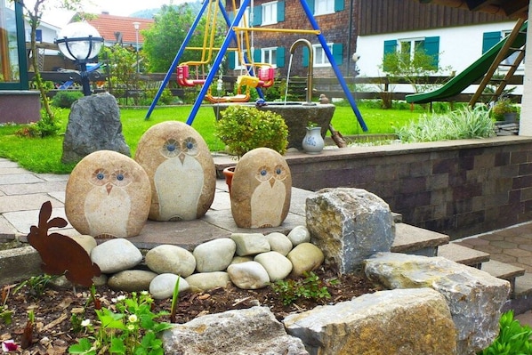 Um jardim com estátuas decorativas de corujas, um parquinho com balanços e escorregadores, e um muro de pedra.