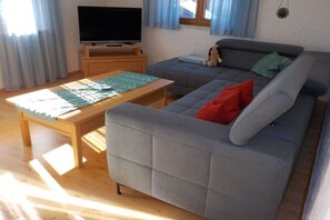 Una sala de estar con un sofá gris, una mesa de centro de madera, un televisor sobre un mueble de madera y una ventana con cortinas.