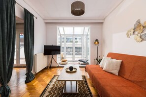 A living room with a sofa, a coffee table, a TV on a stand, and a large window with a view of buildings. - Andromeda: Cozy 2BD apartment in the center with spacious balcony (Thessaloniki)