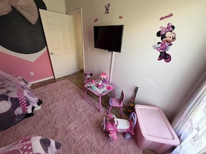 Ein Kinderzimmer mit rosa Teppich, einem kleinen Tisch mit Spielzeug, einem rosa Stuhl, einem Fernseher und Minnie Maus Dekorationen.
