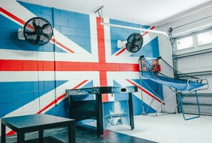 Ein Zimmer mit Wand in den Farben der Union Jack, einem Tisch, einer Bank, einem Ventilator und einem Kunstdruck zum Thema Eishockey.