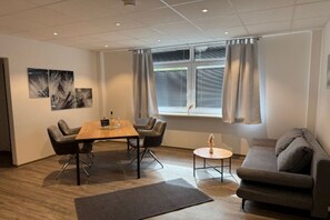 A modern room with a wooden table, two chairs, a grey sofa, a small round table, and a window with curtains. - Holiday apartment with 1 bedroom (Ludwigswinkel)