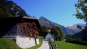 Ein traditionelles Holzchalet mit Steinfundament, eine kleine weiße Kapelle und ein Berg im Hintergrund.