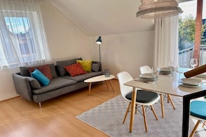 Uma sala de estar moderna com um sofá cinzento, uma mesa de jantar posta para quatro pessoas e uma janela grande com cortinas leves.
