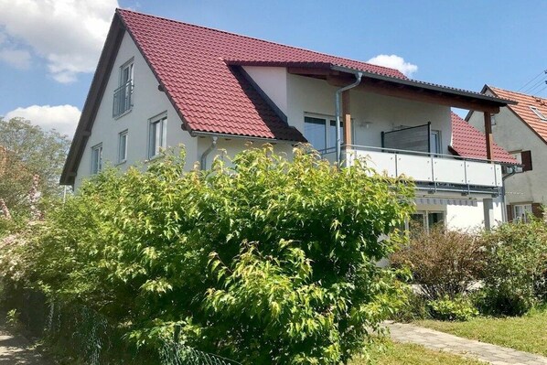 Uma casa de dois andares com telhado de telhas vermelhas, uma varanda e um jardim em primeiro plano.
