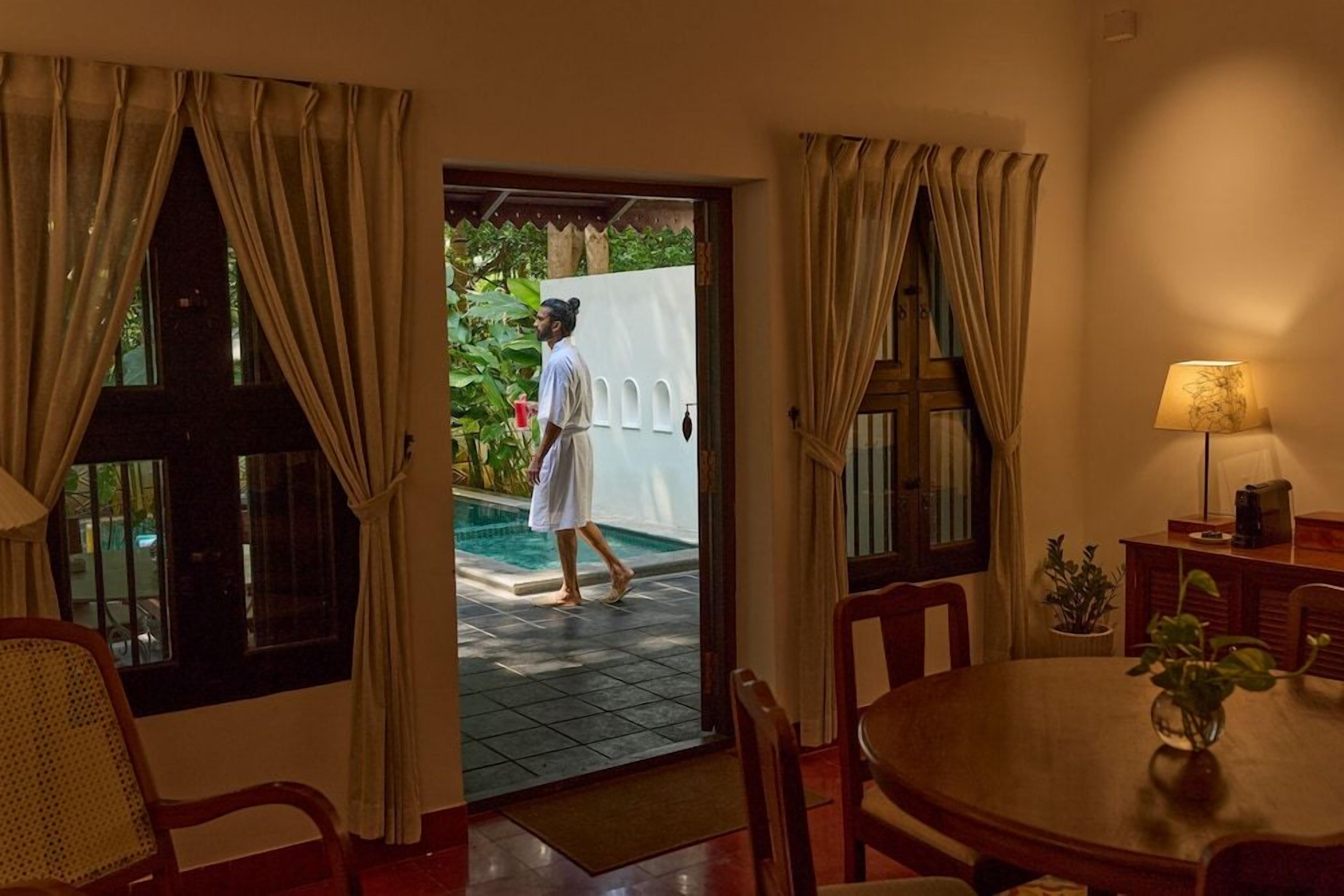 A room with a dining table, chairs, a lamp, and a view of a swimming pool.