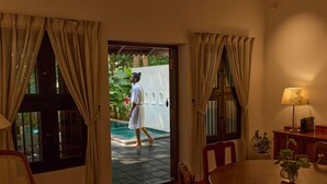 A room with a dining table, chairs, a lamp, and a view of a swimming pool. - Swaswara Wellness - CGH Earth, Gokarna (Kumta)