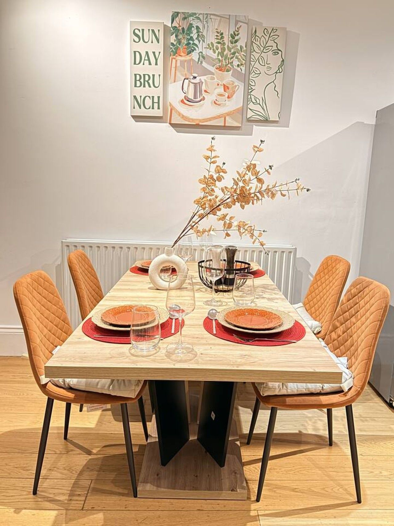 A dining table set for brunch with six chairs, a vase with dried flowers, and a wall art piece above.