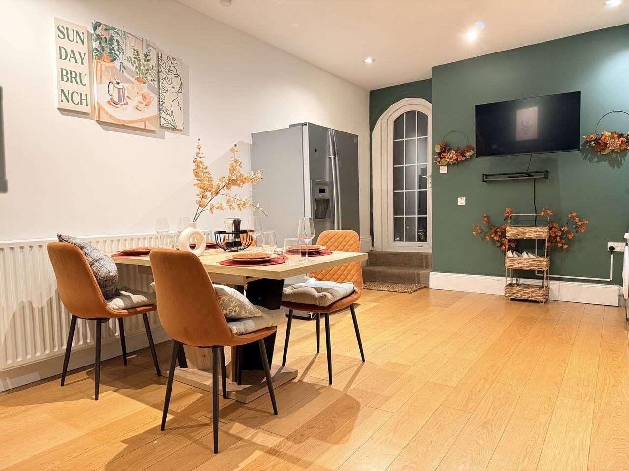 A dining area with a table set for two, chairs, a refrigerator, a television mounted on the wall, and a decorative wall piece.