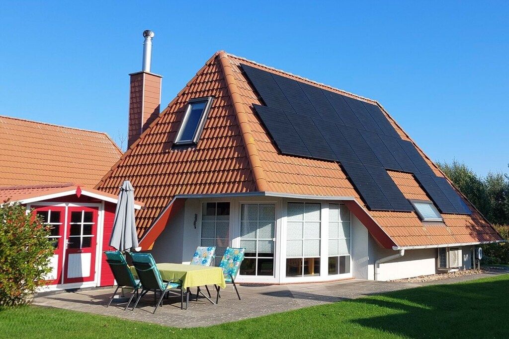 Ein Haus mit geneigtem Dach, Solaranlagen, einem Schornstein und einer Terrasse mit Gartenmöbeln.