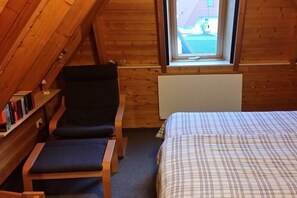 A cozy room with a sloped ceiling, a bed with a plaid bedspread, a dark blue chair, a small table, and a radiator. - Holiday home with 1 bedroom (Wurster Nordseeküste)