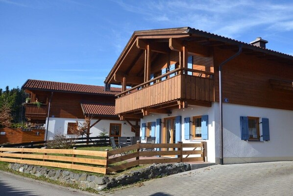 A two-story wooden building with a balcony, a stone wall, and a paved entrance area. - Holiday home in Allgäu with 3 bedrooms (Lechbruck am See)