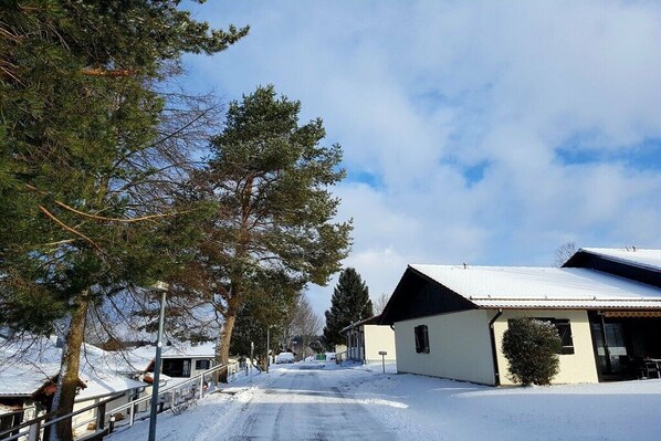 A snow-covered street with buildings, including a house with a sloped roof and a small structure with a flat roof. - Holiday home in Allgäu with 1 bedroom (Lechbruck)