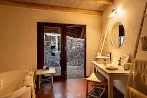A bathroom with a bathtub, a sink, a mirror, and a small table. There is a stone wall visible through an open door. - Zebras Crossing Private Game Lodge (Modimolle)