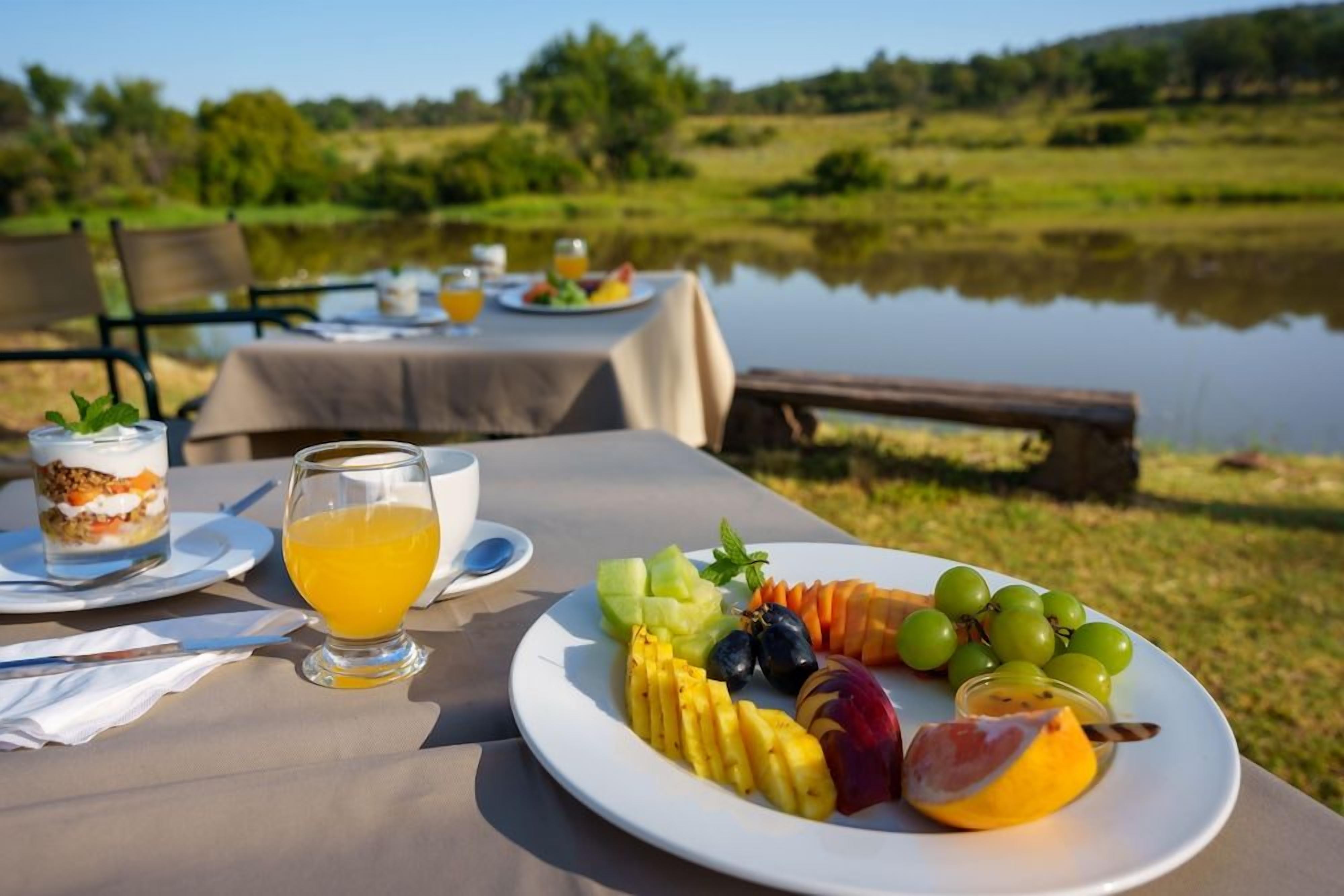 A table set with a fruit platter, a glass of orange juice, and a dessert on a lakeside table.