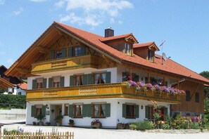 Ein zweistöckiges Gebäude mit rotem Ziegeldach, mehreren Balkonen mit Blumenkästen und einer weißen Fassade mit grünen Fensterläden.