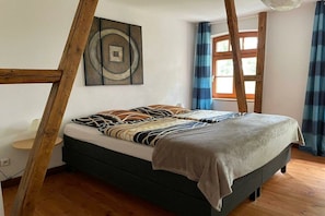 Ein Schlafzimmer mit einem Bett, einer Holzleiter, einem Fenster mit Vorhängen und einem an der Wand befestigten Kunstwerk.
